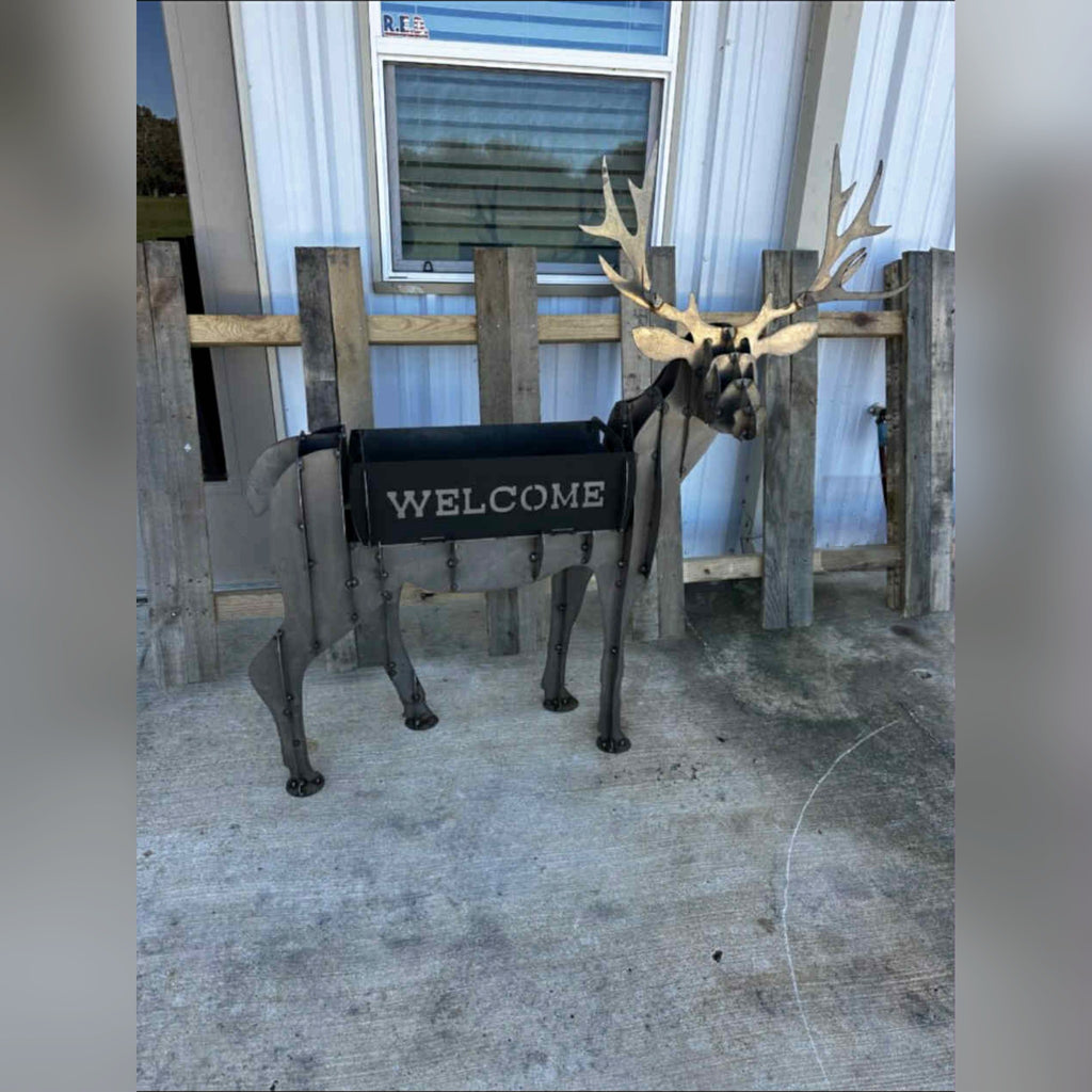 Steel deer planter sculpture with a black WELCOME planter box, in front of a rustic wood fence